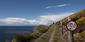 A dirt vehicle track runs along a grassy coastal cliff edge past a round "100 km/h" speed limit sign under a blue sky.