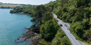 High-angle view of a narrow paved road winding between a lush green forest and a calm turquoise body of water.