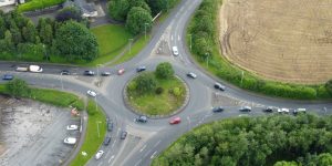 Aerial view of multiple cars navigating a multi-lane roundabout surrounded by green trees and fields.