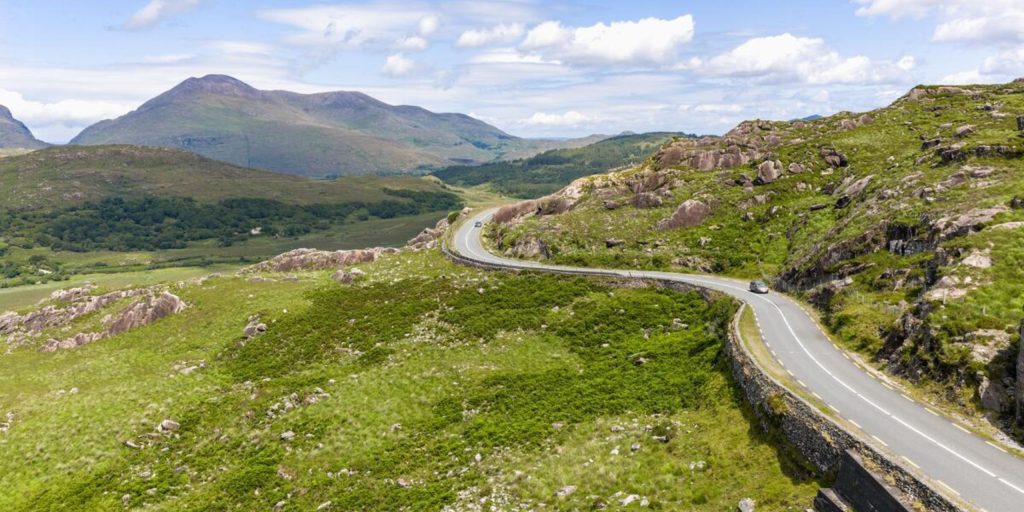A high-angle aerial view of a silver car driving along a curved, winding mountain road that cuts across a vibrant green, grassy hillside under a partly cloudy sky.