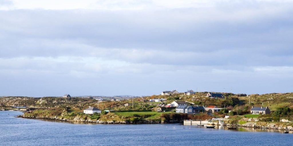 A small coastal village with scattered white and grey houses nestled on low, rocky green hills along a calm blue bay under a bright, overcast sky.