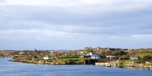 A small coastal village with scattered white and grey houses nestled on low, rocky green hills along a calm blue bay under a bright, overcast sky.
