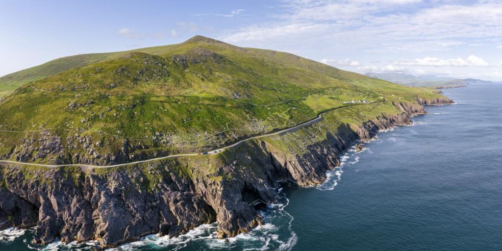 A sweeping aerial shot of a narrow coastal highway carved into the side of a massive, steep green mountain that slopes directly down into rocky ocean cliffs and crashing blue waves.
