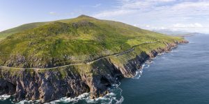 A sweeping aerial shot of a narrow coastal highway carved into the side of a massive, steep green mountain that slopes directly down into rocky ocean cliffs and crashing blue waves.