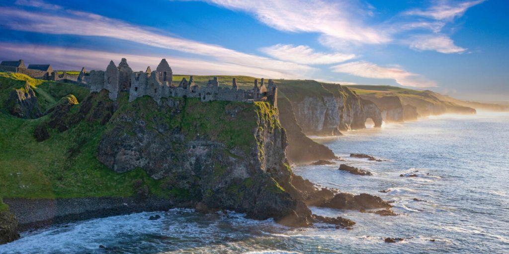 The sprawling, roofless stone ruins of a medieval castle perched precariously on a high coastal cliff, overlooking a sea with crashing white surf and distant, sun-drenched sea stacks.