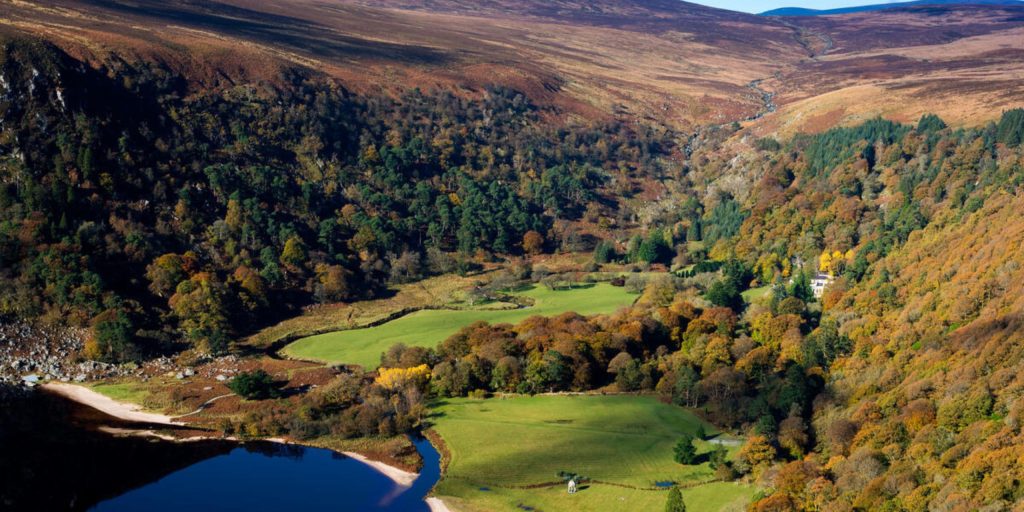 A high-angle view looking down into a deep, lush valley filled with autumn-colored orange and yellow trees, bordered by a dark lake on the left and grassy fields on the valley floor.