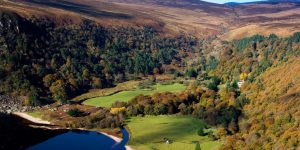 A high-angle view looking down into a deep, lush valley filled with autumn-colored orange and yellow trees, bordered by a dark lake on the left and grassy fields on the valley floor.