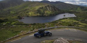 A vintage blue convertible sports car driving around a sharp hairpin curve on a high mountain road, overlooking a large dark lake nestled between rolling green hills below.