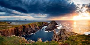 A dramatic sunset over rugged, jagged rock formations jutting out into a deep blue sea, under a sky heavy with dark, swirling storm clouds illuminated by golden light.