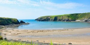 Sandy beach cove sheltered by green, grassy headlands on either side, featuring intricate, swirling patterns drawn directly into the sand near the calm water's edge.