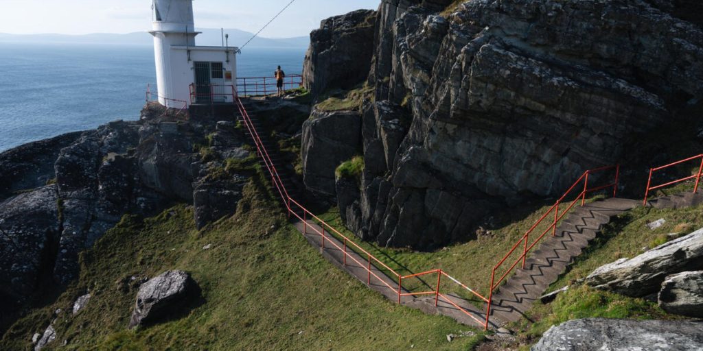 A concrete walkway with bright red metal handrails winding down a steep, rocky cliffside path toward a small white lighthouse tower overlooking a vast blue ocean.