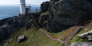 A concrete walkway with bright red metal handrails winding down a steep, rocky cliffside path toward a small white lighthouse tower overlooking a vast blue ocean.