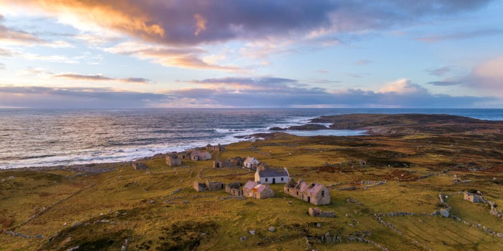 An aerial view at sunset showing the stone ruins of an abandoned coastal village scattered across a grassy, stone-walled landscape next to a choppy sea.