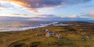 An aerial view at sunset showing the stone ruins of an abandoned coastal village scattered across a grassy, stone-walled landscape next to a choppy sea.