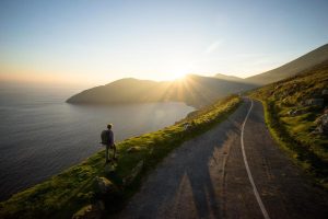 A hiker with a backpack stands on a grassy cliffside overlooking a bay at sunset, next to a winding coastal road.