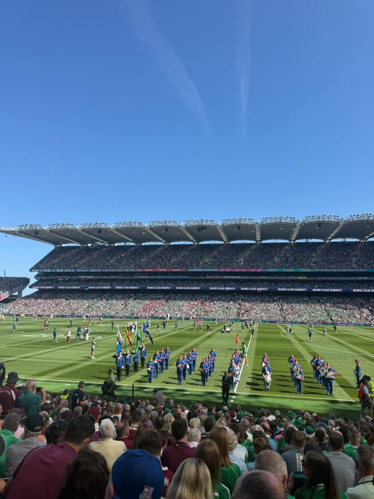 A packed Croke Park stadium under a clear blue sky, showing a marching band in blue and red uniforms performing on the green pitch. A large crowd of spectators in maroon and green jerseys fills the multi-tiered stands, with electronic advertising boards visible in the background.