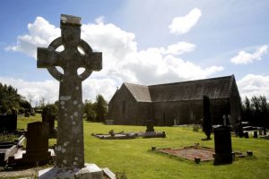 A large Celtic cross stands in the foreground of a peaceful churchyard, with a stone church and gravestones under a bright blue sky with scattered clouds.