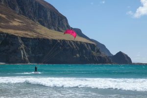 A kitesurfer glides across turquoise water near a sandy beach, with dramatic sea cliffs and rugged mountains rising in the background.