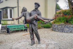 A bronze statue of a man carrying a woman stands in a village square, with a green bench, flower beds, and houses behind it.