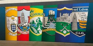 A horizontal wall banner displaying six colorful GAA county crests side by side, including Clare, Cork, Kerry, Limerick, Tipperary and Waterford.