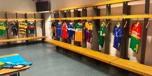 A sports locker room featuring wooden benches and numerous different Gaelic football and hurling jerseys hanging neatly on hooks along the walls.