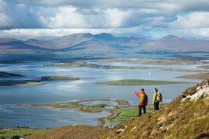 Two hikers stand on a hillside overlooking a vast bay dotted with islands, with mountains and dramatic clouds stretching across the horizon.