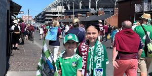 Two young girls smiling for a picture together, in the background is supporters wearing green and maroon.