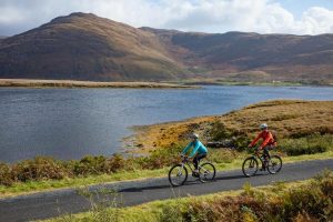 Two cyclists riding along a paved path next to a scenic lake and large hills.