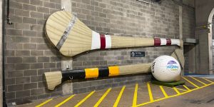 Two massive, oversized decorative hurling sticks mounted horizontally on a grey brick wall next to a large white Gaelic football.