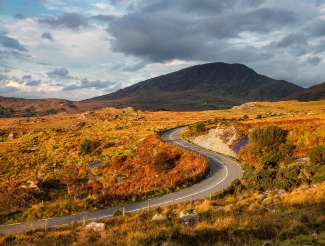 A winding two-lane paved road snakes through a vast mountain pass landscape filled with golden-brown autumnal grasses and low shrubbery under a dramatic, cloudy sky.