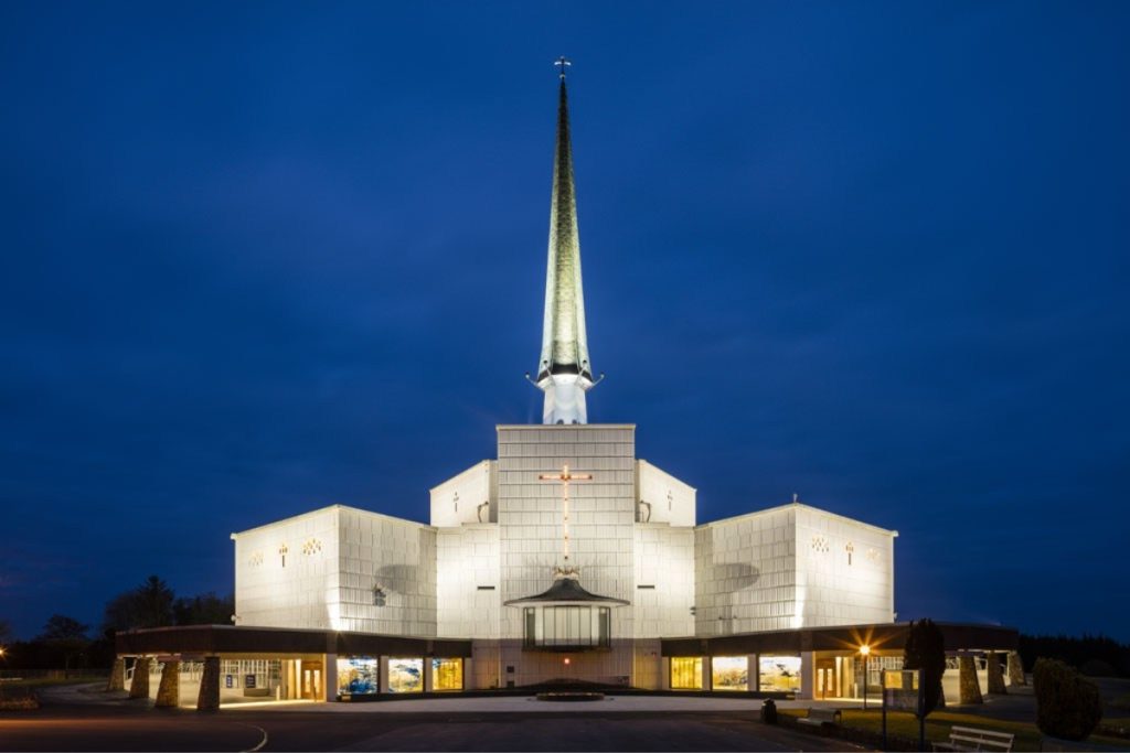 A brightly illuminated modern church with a tall spire stands against a deep blue evening sky, its entrance and façade glowing with warm light.