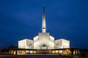 A brightly illuminated modern church with a tall spire stands against a deep blue evening sky, its entrance and façade glowing with warm light.