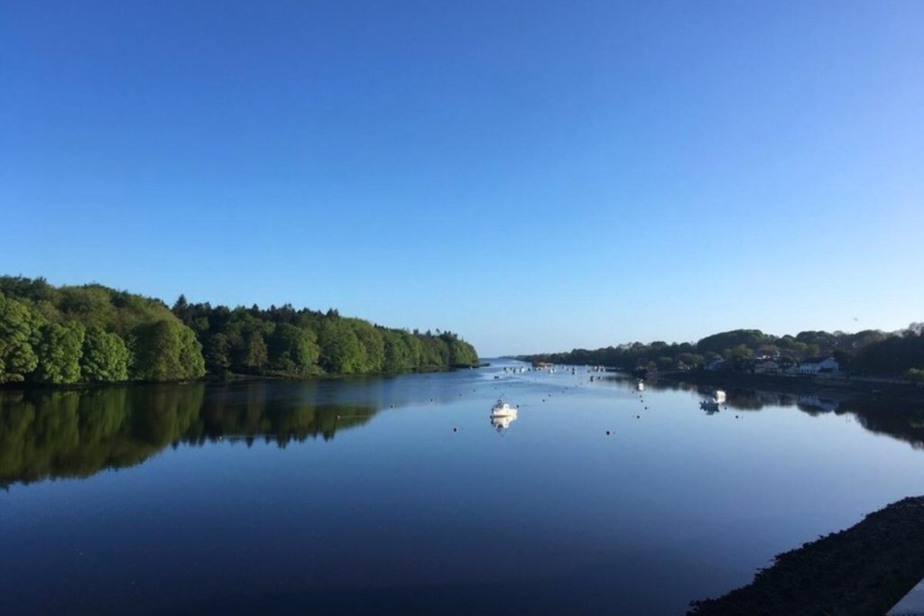 Scenic river with calm blue water reflecting the sky, framed by green trees on the banks, and a few small fishing boats floating peacefully.