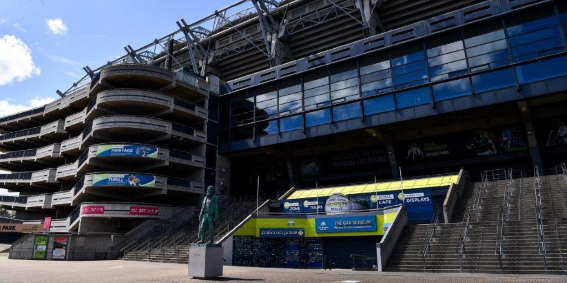 An exterior view of Croke Park stadium in Dublin, showing the large concrete and glass architecture of the stand. A green statue of a man on a pedestal stands in the foreground on a wide paved concourse, leading toward stairs and the entrance of the GAA Museum.