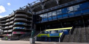 An exterior view of Croke Park stadium in Dublin, showing the large concrete and glass architecture of the stand. A green statue of a man on a pedestal stands in the foreground on a wide paved concourse, leading toward stairs and the entrance of the GAA Museum.