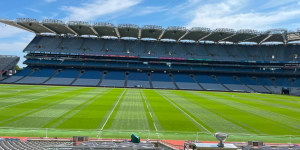 A wide view of an empty Croke Park stadium with blue seating, a distinctive curved roof, and a bright green grass pitch under a blue, partly cloudy sky.