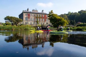 People in canoes paddling on a calm river with a stone castle on the green shore in the background.