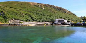 A panoramic view of a calm, turquoise bay with crystal-clear water. A white, two-story stone building sits at the edge of a small harbor beach on the right, backed by a massive, steep green hill covered in lush vegetation.