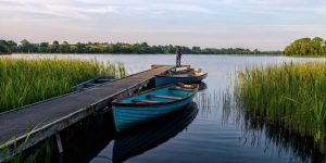 A man fishing from the end of a long wooden pier on a calm lake, with several blue wooden rowboats tied alongside it.