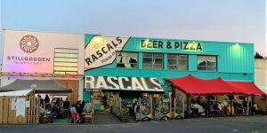 The vibrant exterior of Rascals Brewing Co. and Stillgarden Distillery, featuring teal walls, bold signage, and crowded outdoor patio seating under red awnings.