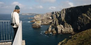 A person wearing a knit sweater and a beanie stands on a metal viewing platform, looking out at dramatic, layered rock cliffs jutting into a deep blue ocean under a partly cloudy sky.