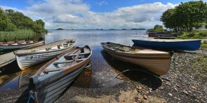 Several traditional wooden rowboats parked along a rocky, pebbled lakeshore under a bright, partly cloudy sky.
