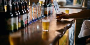 A hand reaching for a small glass of beer on a long, polished wooden bar counter lined with beer bottles and glass awards.