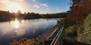 Bright sunlight flare over a wide river, viewed from an elevated wooden dock structure with metal railings.