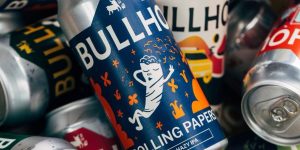 A close-up pile of aluminum beer cans, focusing on a blue Bullhouse Brew Co. "Rolling Papers" hazy IPA can with an illustrated white character.