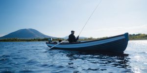 An older man casting a fishing line from a white and blue rowboat on open water, with a large rounded mountain in the background.