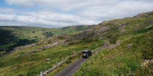 A dark off-road vehicle drives along a narrow, winding asphalt road cutting through a rugged, green mountain landscape filled with rocky outcrops and stone walls.