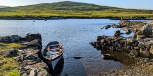 A single rowboat moored in a small, rocky inlet on a clear lake, with a large green hill rising up across the water.