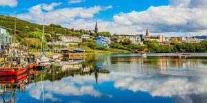 A scenic waterfront view of a coastal Irish town shows colorful boats moored along a calm bay, with vibrant buildings and a church spire reflecting perfectly in the still water.