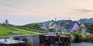 An outdoor patio dining area set with wooden tables and glasses overlooks a rolling green landscape and a quaint village street lined with traditional cottages at dusk.